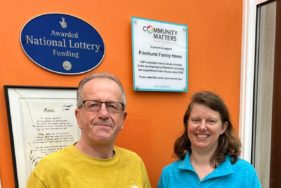 CEO of Community Matters Partnership and Frimhurst Family House team member pose under a plaque to celebrate their partnership.