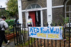 Two people are entering a house, and a banner pointing to the door reads: "ATD Fourth World, Openb House". There are balloons near the banner and bunting above the door.