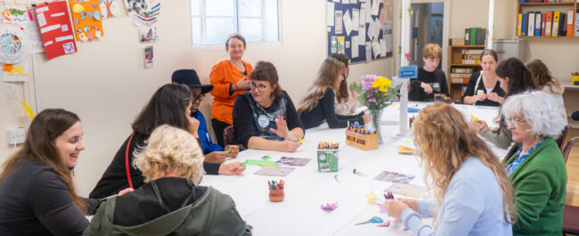 In a room with a big table at the centre, many adults are sat and colouring. Some are chatting, sopme are smiling. There are also some doing origami.