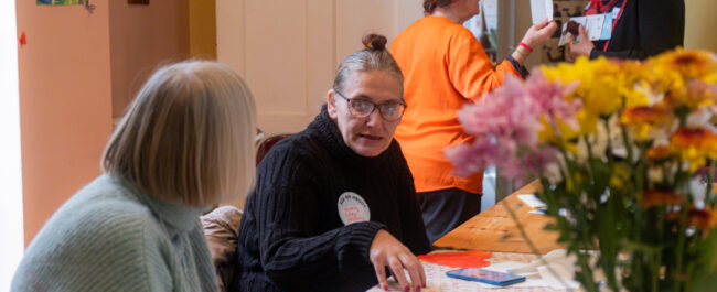 Two women are at a table, one is speaking and the other is attentively listening. On the table are drawings, and colourful flowers. In the background, two women are chatting.