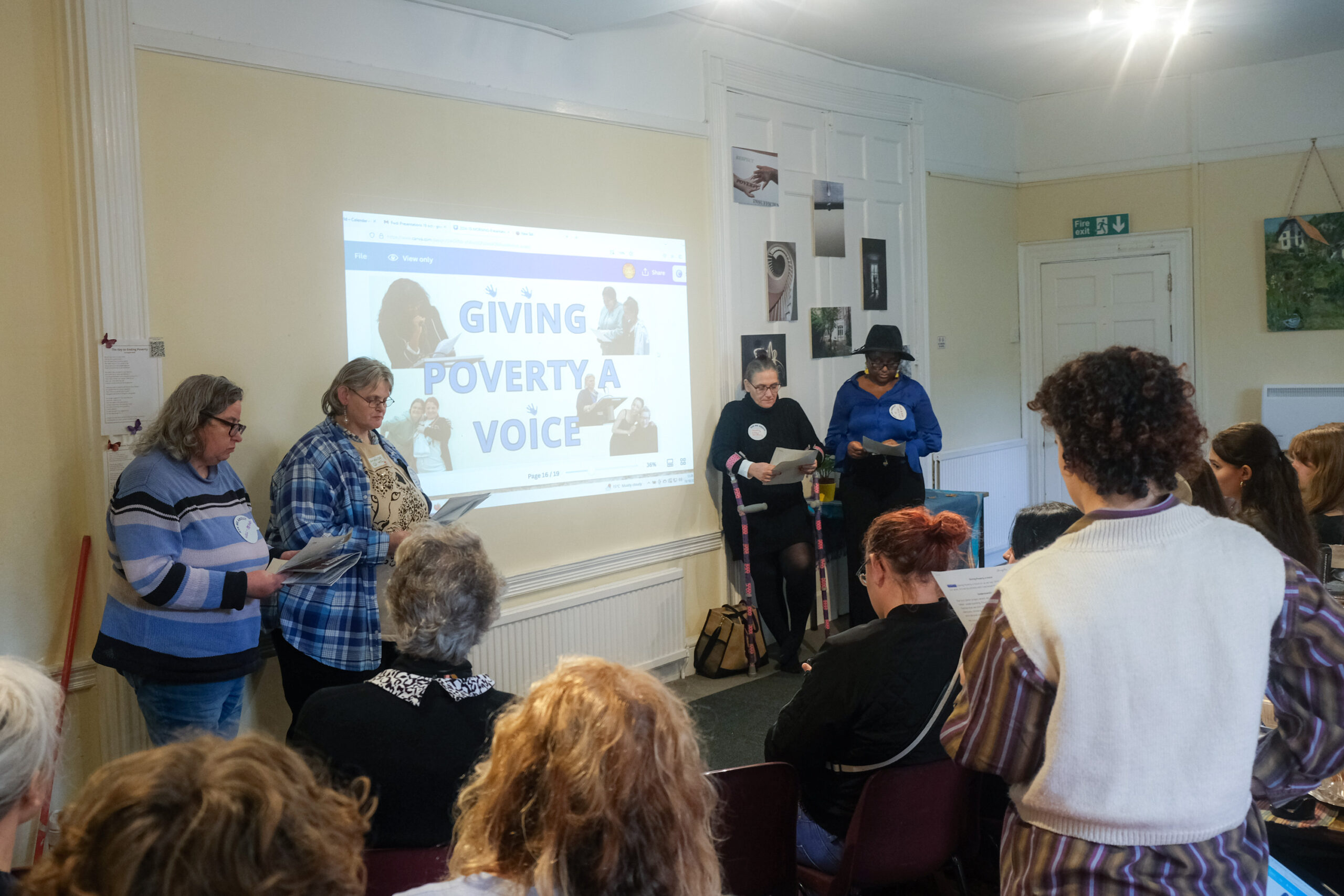 Four women are standing in front of a wall where a screen is projected. The screen reads "Giving Poverty A Voice". In the foreground, people are sat and listening to the women. 