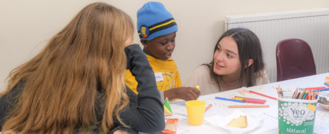 A table is covered in white paper and filled with drawings. Two women are at either side of a little boy, who is drawing and smiling.