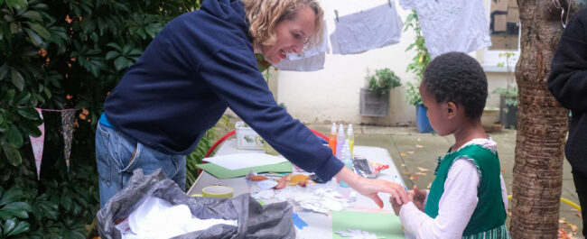 At a table, a woman is helping a little girl do an arts and crafts activity. On the table are papers, pens, leaves and paint. It is in a courtyard, there is a tree behind, and a string where some decorated tee-shirts are hung.