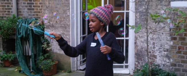 In a courtyard, a little girl is holding a stick to make bubbles. She is surrounded by bubbles and she is smiling.