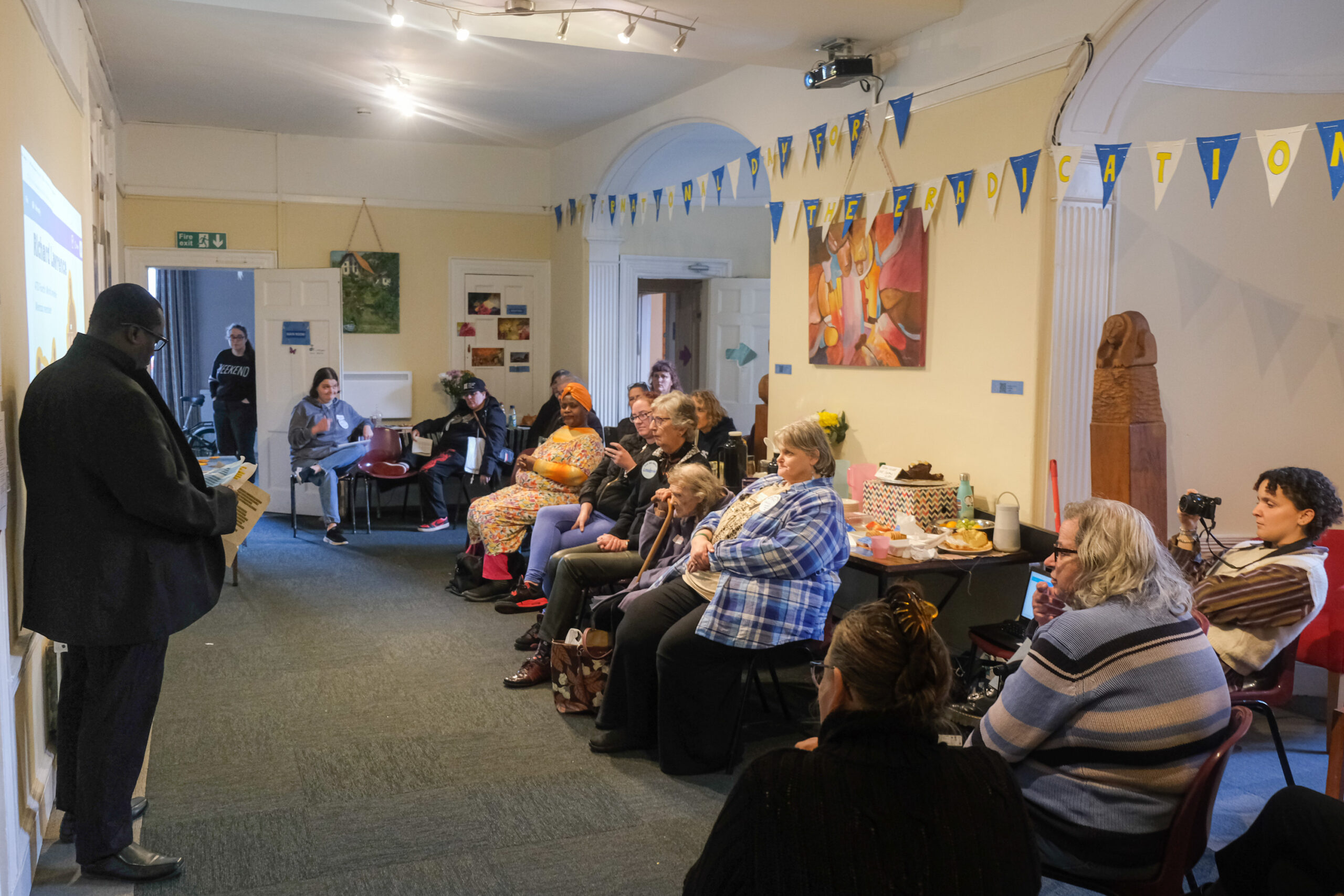 A man, on the left, is standing and reading from a paper. He speaks in front of an audience of about 10 people, sat on the right. The room is decorated with paintings and bunting.