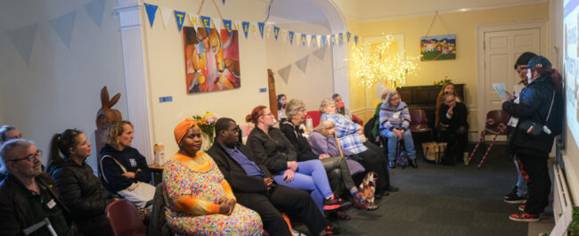 Many adults are sat on the left of a room, and looking at three people standing and speaking on the right. The room is decorated with bunting and paintings.
