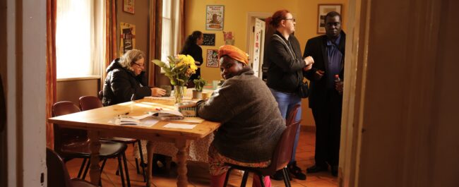 Five adults are in a room, two are chatting, the others are around a table with a cup of tea.