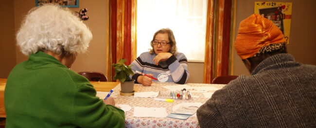 Three women at a table, two have their backs turned to the camera, they are drawing. One woman faces the camera, at the centre, she is holding a pen and looking in the distance.