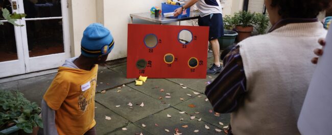 A child and an adult are in the foreground, their back turned to the camera, and looking at a big wood board where there are holes and numbers painted. Bean bages are at the foot of a board. A third person, a man, is picking up chalk from a table nearby.