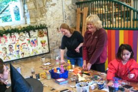 Two women are standing around a table, and othersare seating. There is also a child doing a craft activity with glue. The women are laughing. The table is filled with craft supplies.