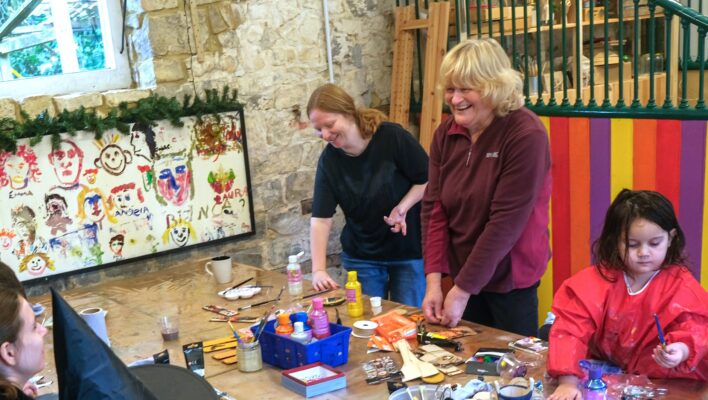 Two women are standing around a table, and othersare seating. There is also a child doing a craft activity with glue. The women are laughing. The table is filled with craft supplies.