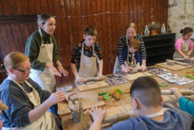 Around a table, children and adults are making pottery: they are rolling clar, while a woman is showing them how to do it.