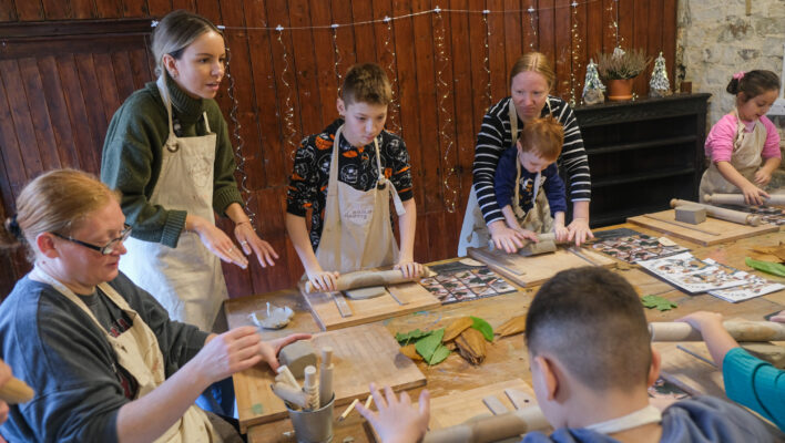 Around a table, children and adults are making pottery: they are rolling clar, while a woman is showing them how to do it.